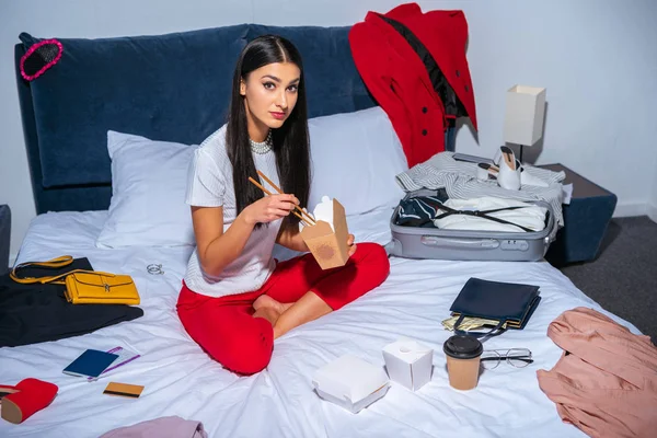 High angle view of attractive brunette girl eating asian food and looking at camera while sitting on bed with suitcase and clothes — Stock Photo