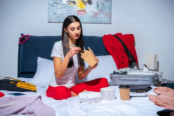 Atractiva chica morena comiendo comida asiática mientras está sentado en la cama con maleta y ropa - foto de stock