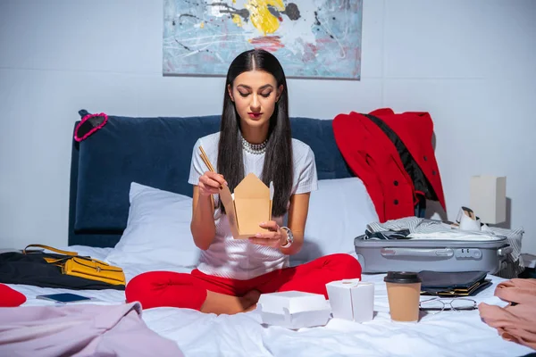 Hermosa chica morena sentada en la cama y comer comida para llevar antes del viaje - foto de stock