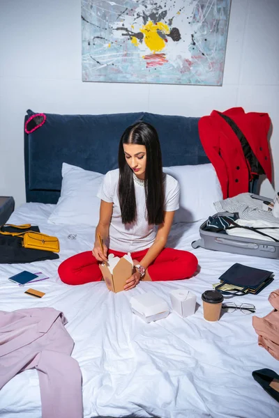 Vista ad alto angolo di bella ragazza seduta sul letto e mangiare cibo da asporto prima del viaggio — Foto stock