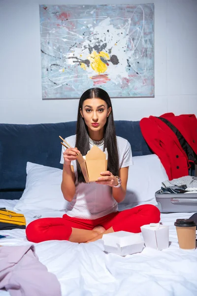 Приваблива молода жінка їсть забирає їжу і дивиться на камеру, сидячи на ліжку — стокове фото