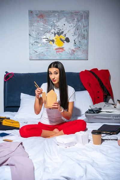Beautiful young woman eating take away food and looking at camera while sitting on bed — Stock Photo