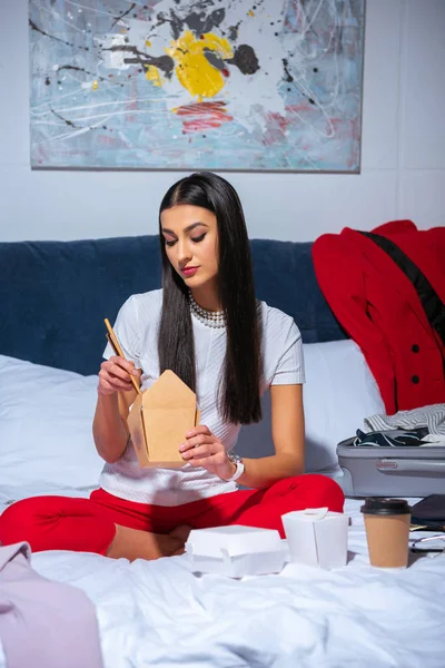 Молода брюнетка сидить на ліжку і їсть азіатську їжу — стокове фото