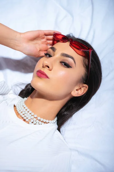 Jolie femme brune ajustant les lunettes de soleil rouges et regardant la caméra tout en étant couché sur le lit — Photo de stock