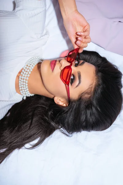 High angle view of elegant brunette girl in red eyeglasses lying on bed and looking at camera — Stock Photo