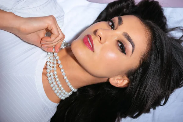 Beautiful woman in pearl necklace lying on bed and looking at camera — Stock Photo