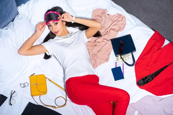 High angle view of young woman in eye mask looking at camera while lying on bed with stylish clothes, passport, boarding pass and dollar banknotes — Stock Photo