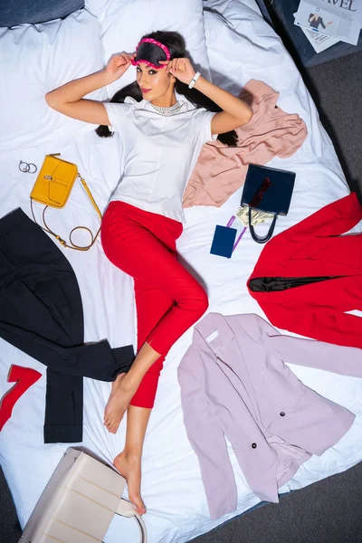 Draufsicht einer jungen Frau mit Augenmaske, die mit stylischer Kleidung, Pass, Bordkarte und Dollarnoten im Bett in die Kamera blickt — Stockfoto
