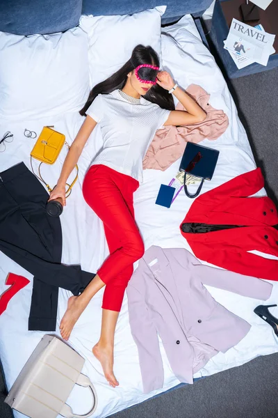 Вид сверху на молодую женщину в маске для глаз спящую на кровати с кофе на вынос, паспорт и посадочный талон — стоковое фото
