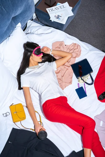 Vista de ángulo alto de la mujer joven en máscara para los ojos durmiendo en la cama con café para llevar, pasaporte y tarjeta de embarque - foto de stock