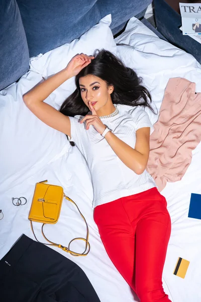 High angle view of young woman with finger on lips looking at camera while lying on bed — Stock Photo