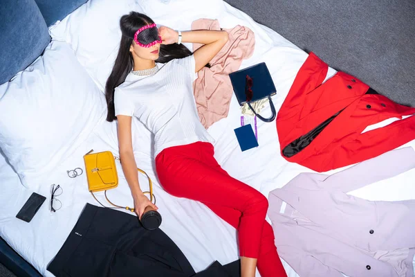 Vista superior da menina em máscara olho segurando café para ir e dormir na cama com roupas, passaporte e dinheiro — Fotografia de Stock