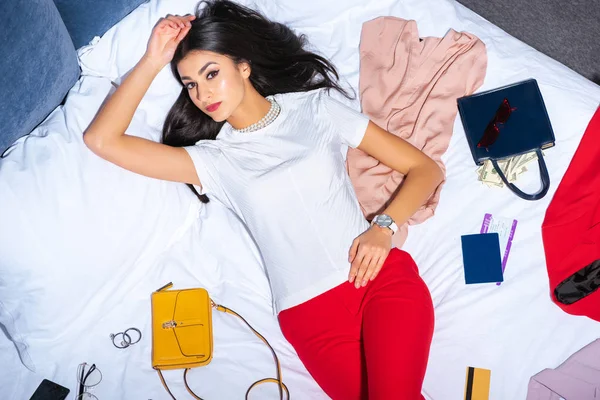 Top view of beautiful girl looking at camera while lying on bed with fashionable clothes, handbags with money, credit card and passport with ticket — Stock Photo
