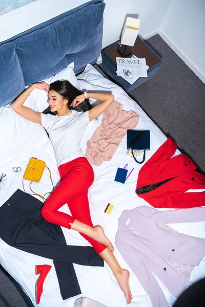 High angle view of happy girl lying on bed with fashionable clothes, credit card, passport and travel newspaper — Stock Photo