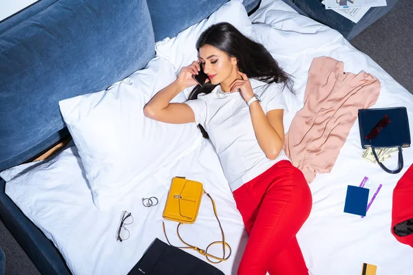 Vista de ángulo alto de hermosa chica sonriente hablando por teléfono inteligente mientras está acostado en la cama con ropa elegante, pasaporte y tarjeta de embarque - foto de stock