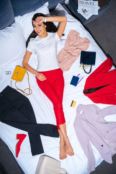 Belle jeune femme souriant à la caméra tout en étant couché sur le lit avec des vêtements à la mode, passeport, carte de crédit et journal de voyage — Photo de stock