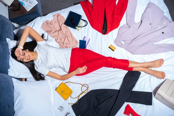 Draufsicht auf eine lächelnde junge Frau, die mit verschiedenen stylischen Kleidungsstücken, Handtaschen und Kreditkarte im Bett liegt — Stockfoto