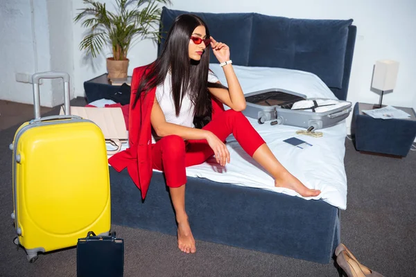 Vista de ángulo alto de la hermosa mujer joven en traje rojo de moda y gafas de sol sentados en la cama y mirando hacia otro lado - foto de stock