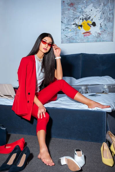 Bella giovane donna in abito rosso alla moda e occhiali da sole seduti sul letto e guardando la fotocamera — Foto stock