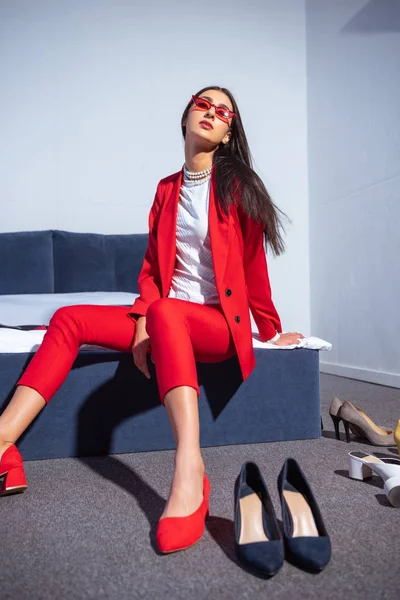 Attractive young woman in fashionable red suit and sunglasses sitting on bed — Stock Photo