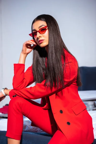 Belle jeune femme en costume rouge élégant et lunettes de soleil assis et regardant la caméra — Photo de stock