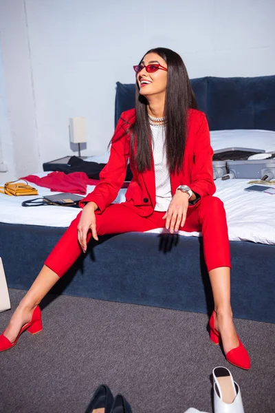 Belle jeune femme souriante en costume rouge élégant et lunettes de soleil assis sur le lit — Photo de stock
