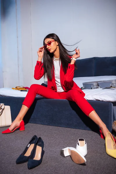 Attraktive junge Frau im schicken roten Anzug und Sonnenbrille auf dem Bett sitzend — Stockfoto