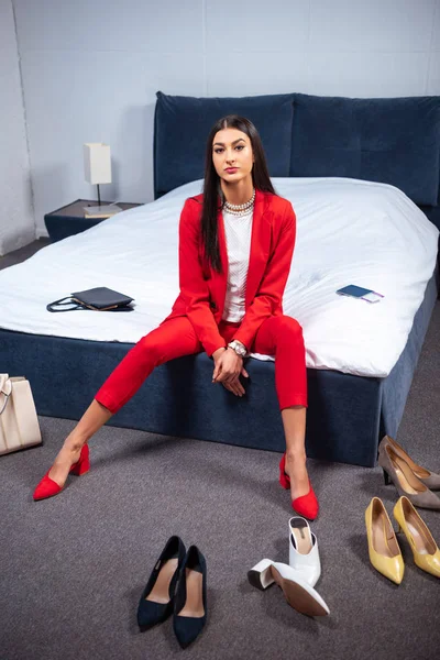 Bella giovane donna in abito rosso elegante seduto sul letto e guardando la fotocamera, varie scarpe alla moda sul pavimento — Foto stock