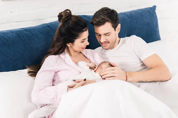 Retrato de mãe sorridente amamentando bebê com o marido perto na cama em casa — Fotografia de Stock