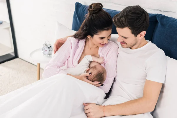 Portrait of smiling mother breastfeeding little baby with husband near by on bed at home — Stock Photo
