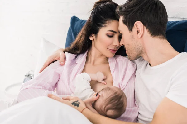 Portrait of young mother breastfeeding little baby with husband near by on bed at home — Stock Photo