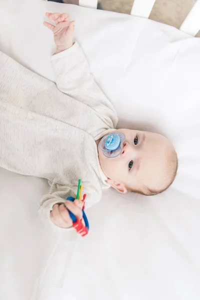 Overhead view of little baby with pacifier holding toy while lying in crib — Stock Photo