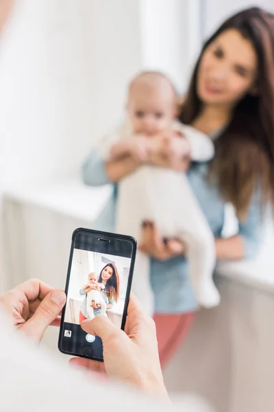 Cropped shot of man taking picture of wife with baby in hands on smartphone — Stock Photo