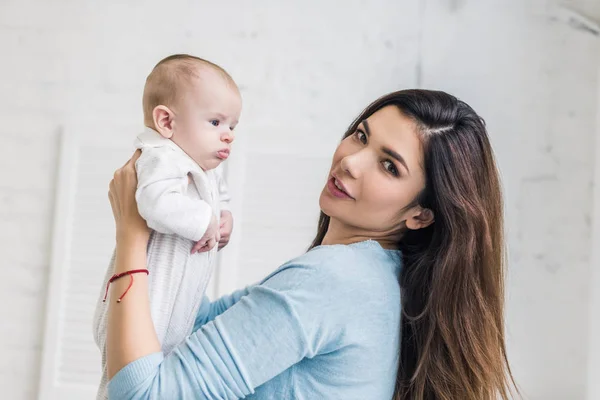 Seitenansicht einer jungen Mutter, die in die Kamera schaut, während sie ihr Baby in den Händen hält — Stockfoto