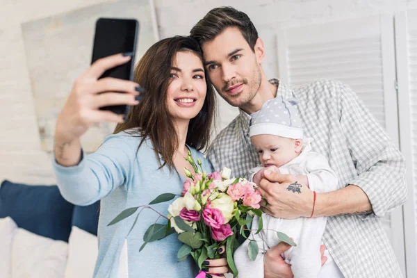 Portrait of smiling woman taking selfie together with husband, little baby and bouquet of flowers on smartphone at home — Stock Photo