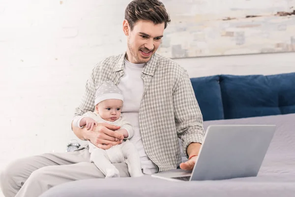 Portrait of man working on laptop with little baby in hands at home, work and life balance concept — Stock Photo