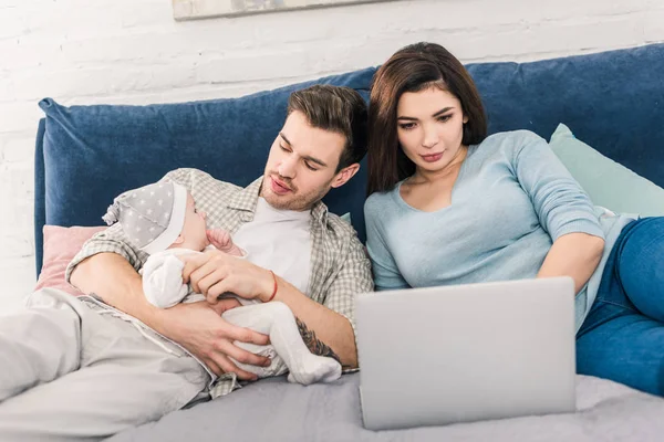 Mujer joven usando el ordenador portátil, mientras que el marido pasar tiempo con su hijo pequeño cerca de - foto de stock