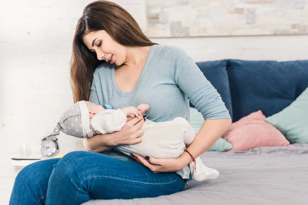 Retrato de madre joven con bebé pequeño con chupete sentado en la cama en casa - foto de stock