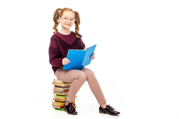 Adorável estudante em óculos sentado em livros e sorrindo para a câmera isolada no branco — Fotografia de Stock