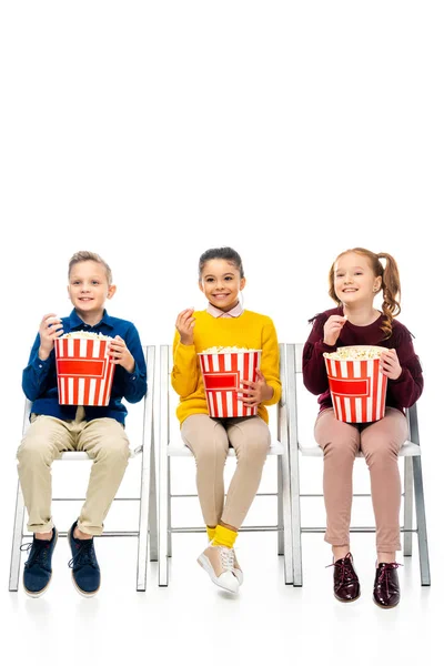 Niños alegres sentados en sillas, sosteniendo cubos a rayas y comiendo palomitas de maíz aisladas en blanco - foto de stock