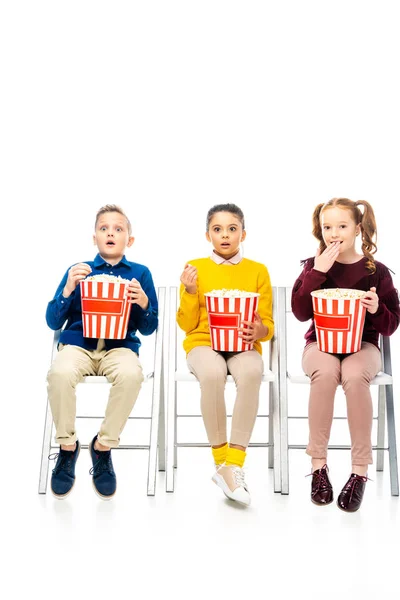 Amazed kids sitting on chairs, holding striped buckets, watching movie and eating popcorn isolated on white — Stock Photo