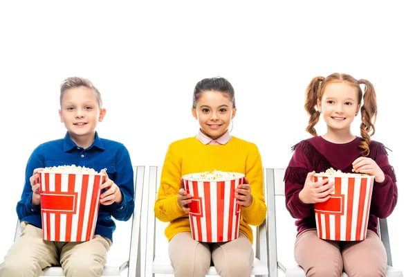 Niños lindos sentados en sillas, sosteniendo cubos de palomitas de maíz a rayas y mirando a la cámara aislada en blanco - foto de stock