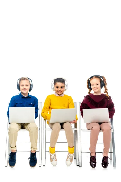 Cute children with headphones sitting on chairs, holding laptops and looking at camera isolated on white — Stock Photo