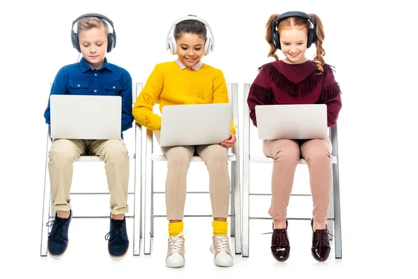 Niños lindos con auriculares sentados en sillas, sonriendo y usando computadoras portátiles aisladas en blanco - foto de stock