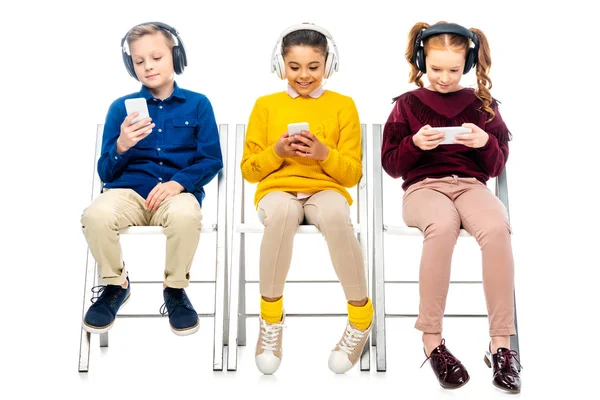 Милі діти з навушниками, сидячи на стільцях і використовуючи смартфони ізольовані на білому — стокове фото