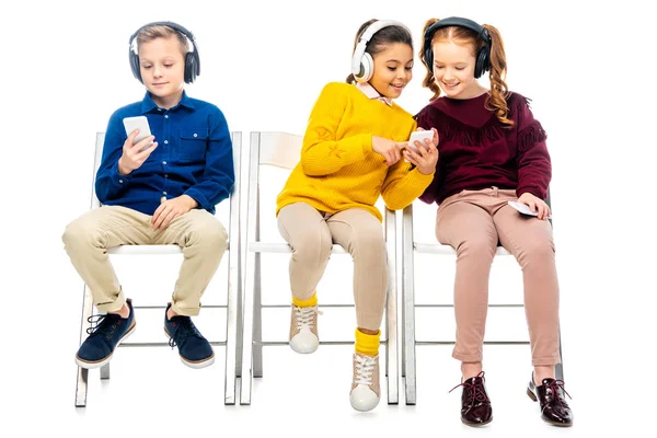 Cute schoolgirls and boy with headphones looking at smartphone screens isolated on white — Stock Photo