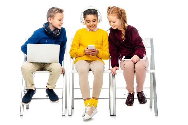 Niños lindos sosteniendo dispositivos digitales y mirando la pantalla del teléfono inteligente de la colegiala linda con auriculares en la cabeza aislada en blanco - foto de stock