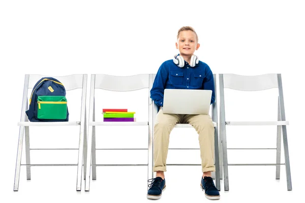 Милий хлопчик з навушниками на шиї сидить на стільці, тримає ноутбук і дивиться на камеру ізольовано на білому — стокове фото