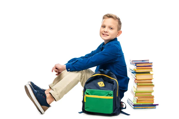 Smiling boy looking at camera and sitting near backpack and stack of books isolated on white — Stock Photo