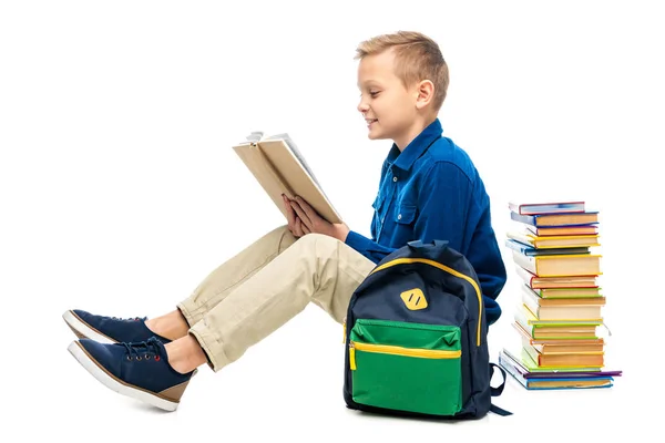 Niño sonriente leyendo libro y sentado cerca de la mochila aislado en blanco - foto de stock